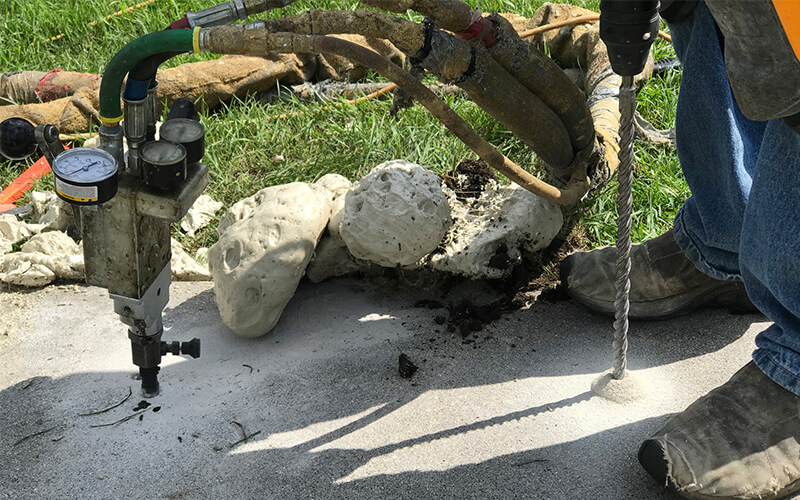 Worker drilling into concrete with a gauge and hoses attached to machinery.