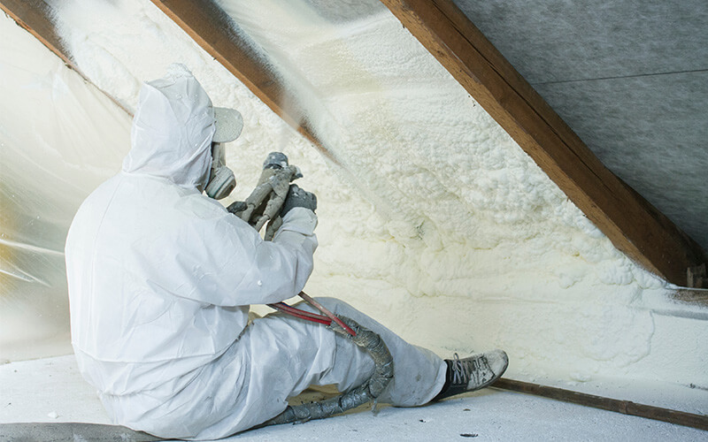 Worker in protective gear applying foam insulation in attic space.